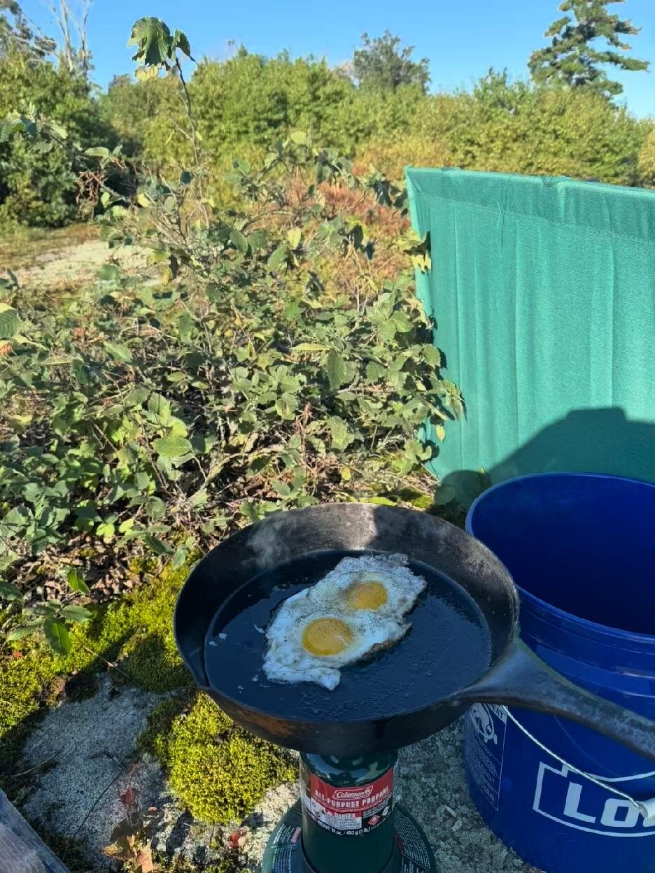 Two fried eggs in a cast-iron pan on a propane burner beside a blue bucket