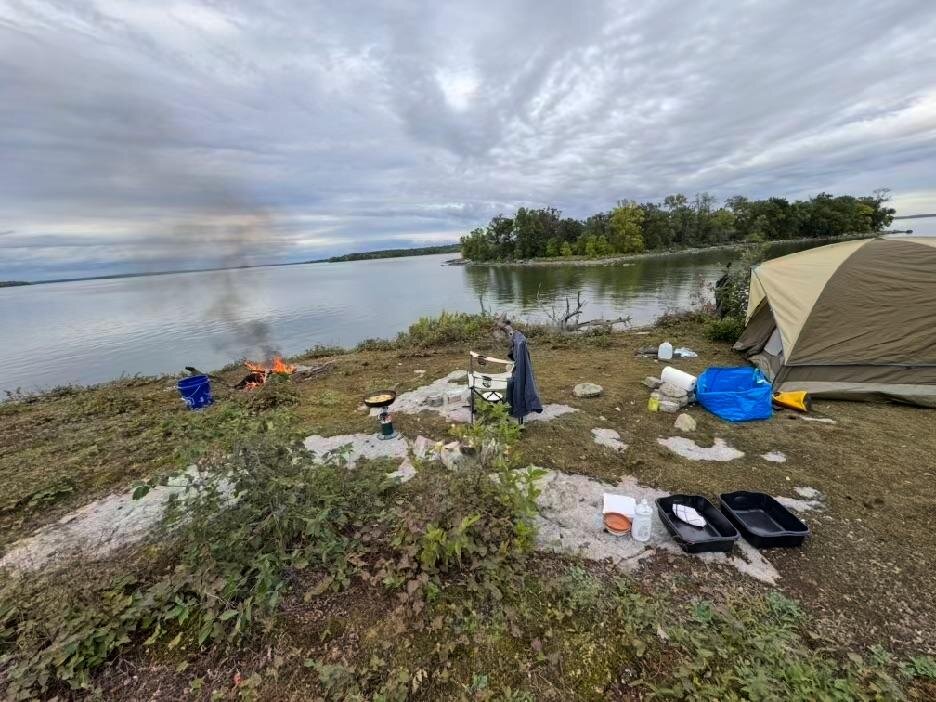 Campfire burning beside a pitched tent on a low rock shoreline, with gear and wash tubs laid out on the moss