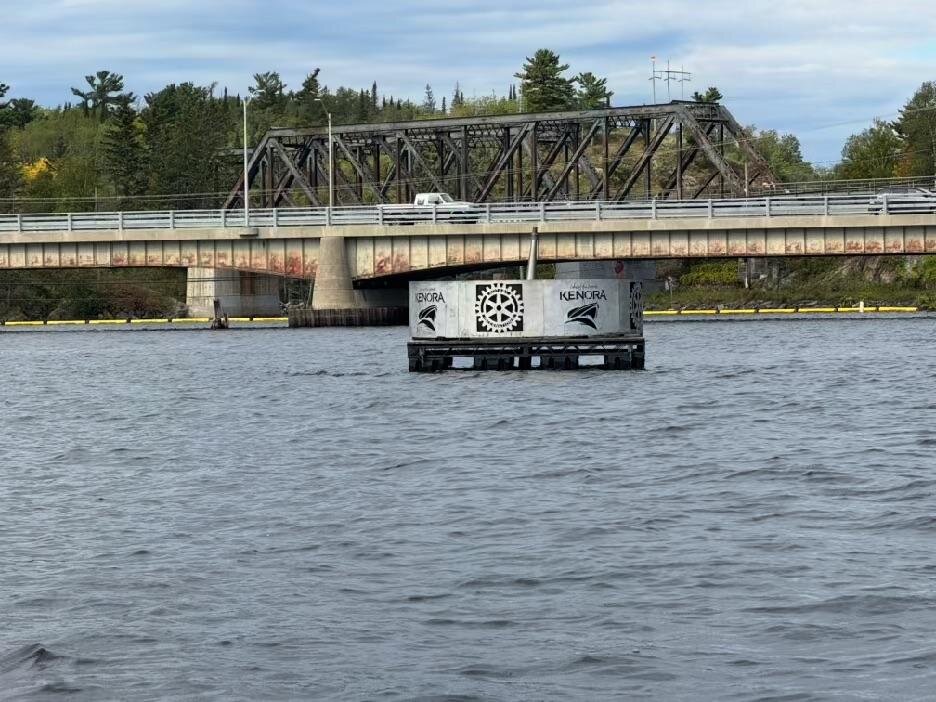 A floating concrete marker with Kenora logos in front of a highway bridge and a steel truss rail bridge