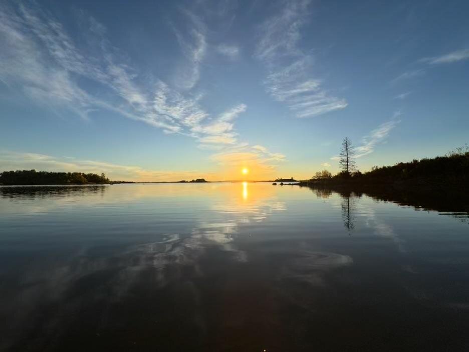 The sun setting over calm water, with thin clouds reflected across the surface
