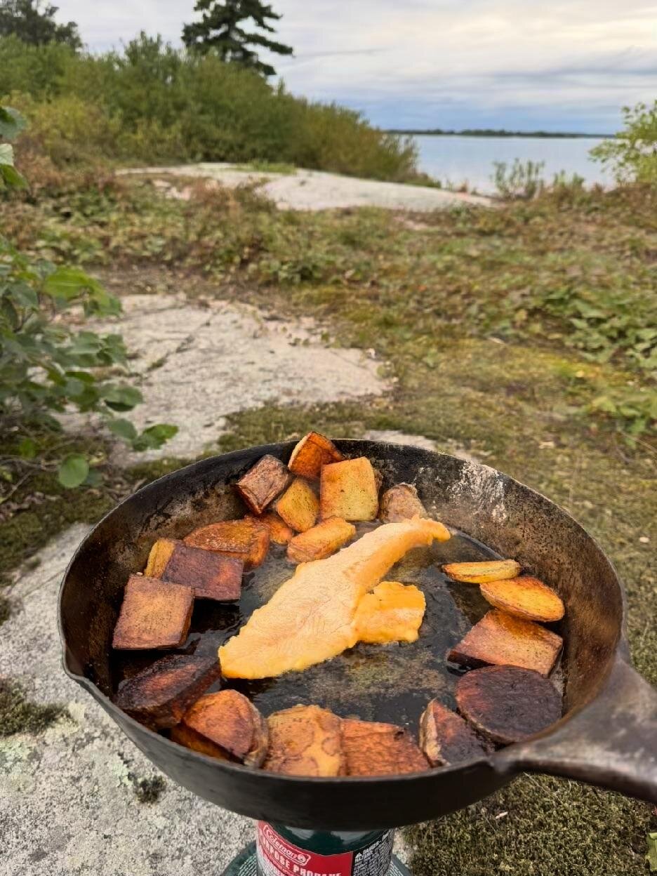 Walleye fillet and sliced potatoes frying in a cast-iron pan over a propane burner, with the lake behind