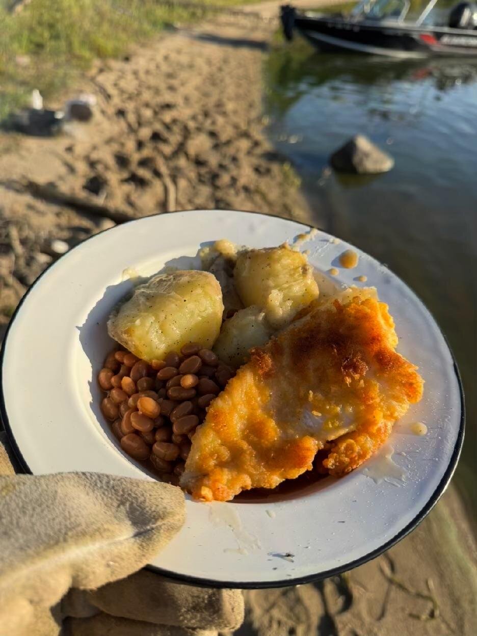 An enamel plate of breaded fried walleye, boiled potatoes, and baked beans, held up in front of the beach