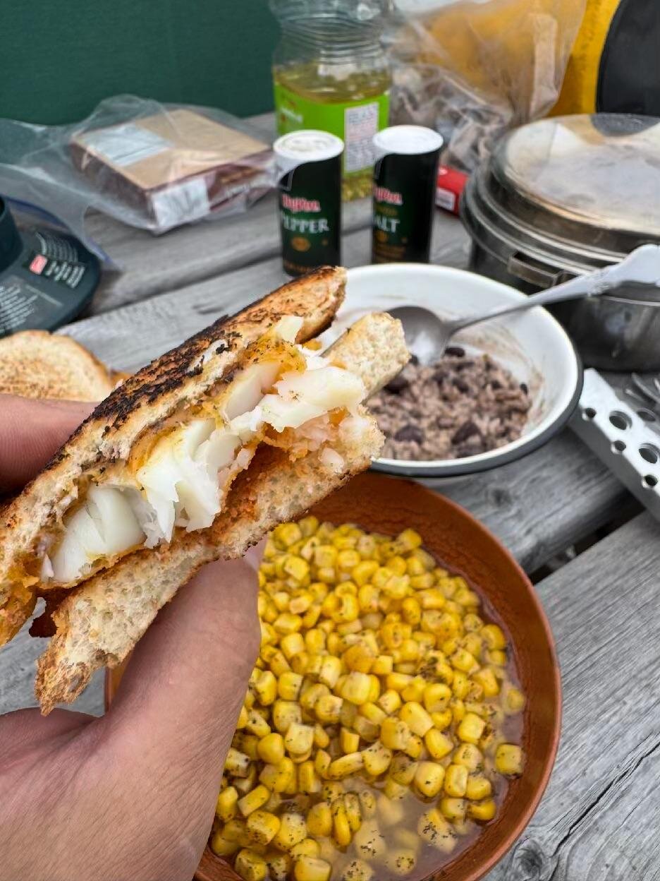 A walleye sandwich held up over bowls of sweet corn and beans and rice on a picnic table